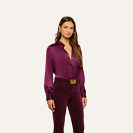 Wearing the Bianca Silk Blouse in deep purple with matching velvet pants, a woman stands against a plain light background, gazing at the camera with a neutral expression.