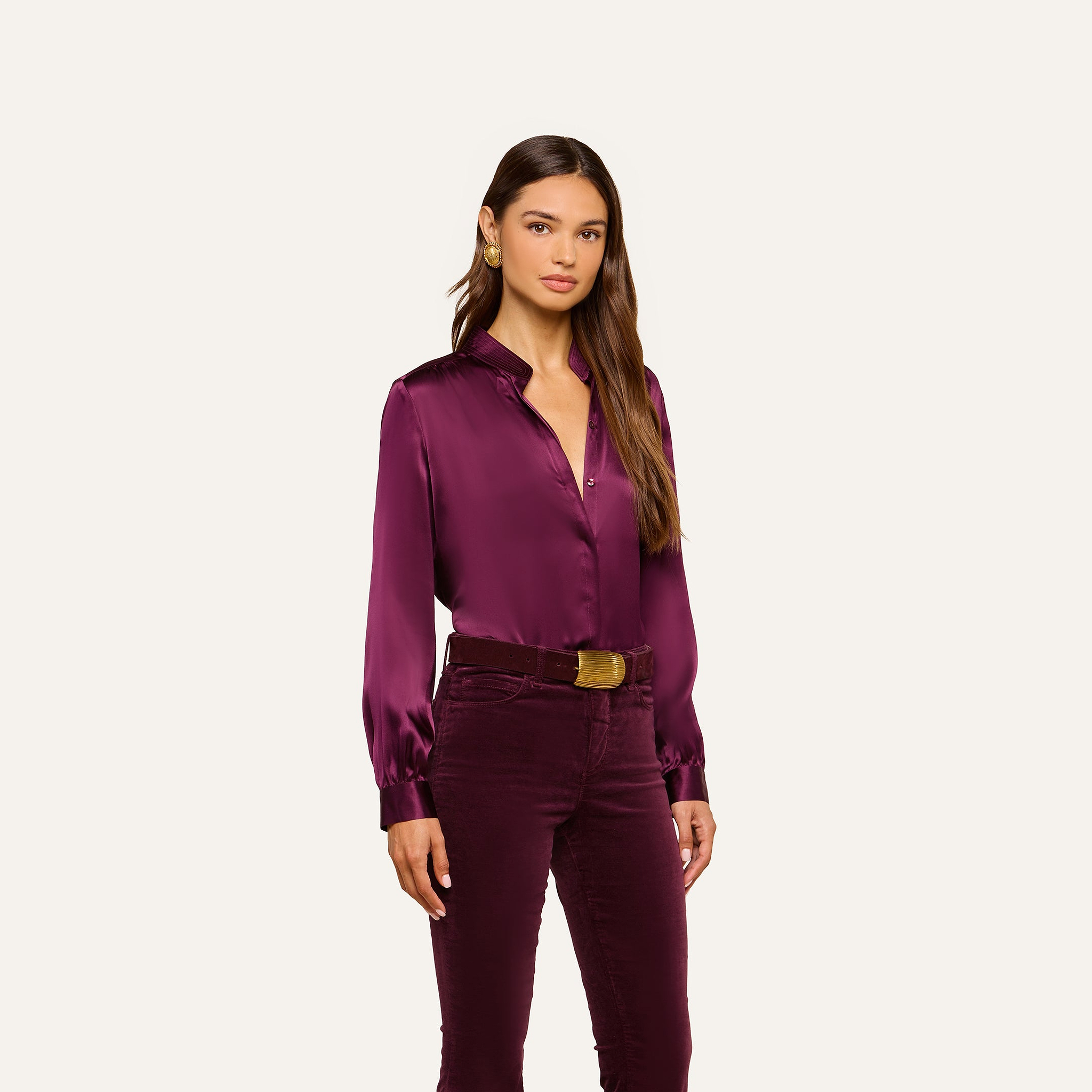 Wearing the Bianca Silk Blouse in deep purple with matching velvet pants, a woman stands against a plain light background, gazing at the camera with a neutral expression.
