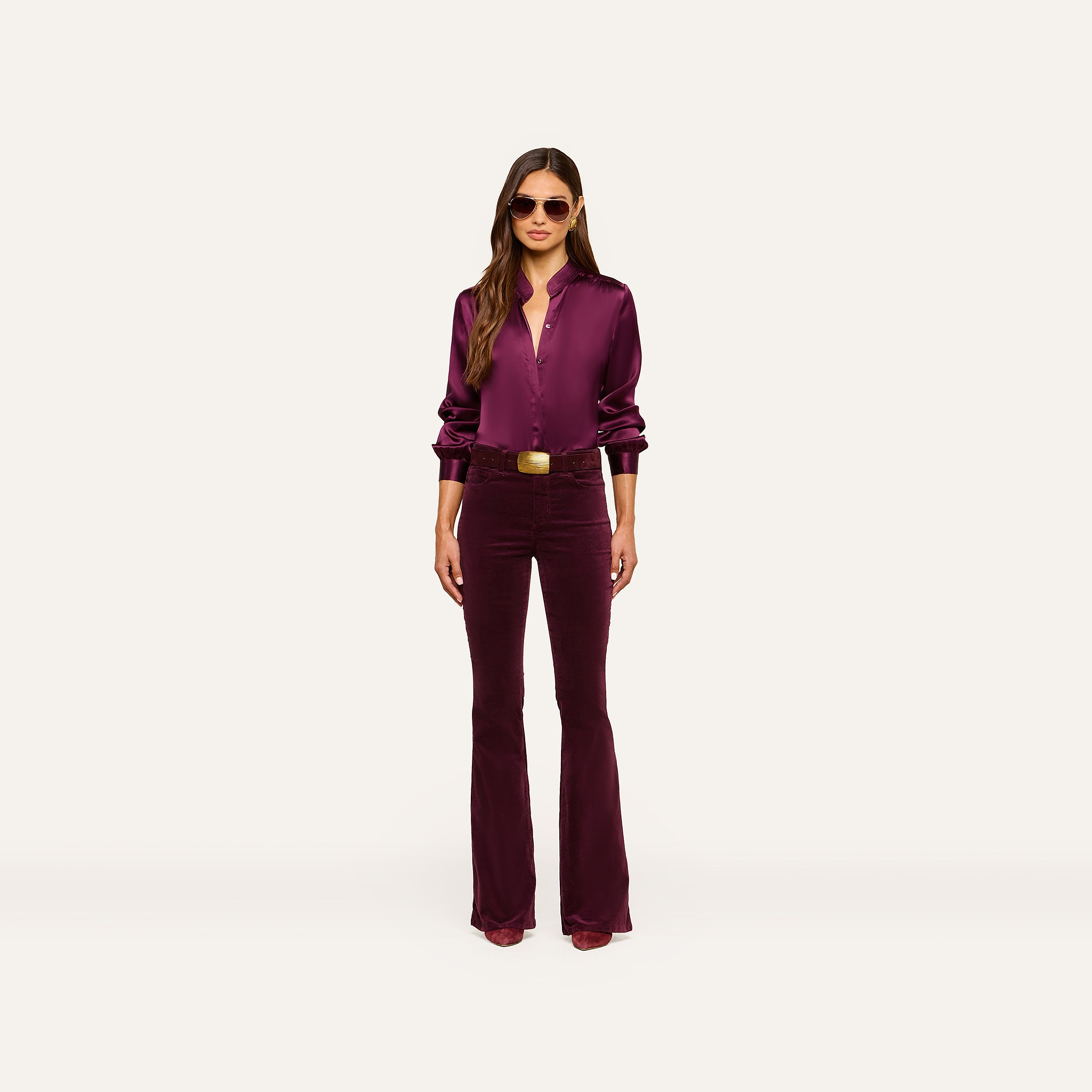 A woman in the Bianca Silk Blouse—crafted from burgundy silk charmeuse—paired with matching flared pants, sunglasses, and heeled shoes stands against a plain white background.
