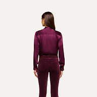 A woman stands facing away, wearing the Bianca Silk Blouse in deep purple with matching velvet pants, against a plain light background.