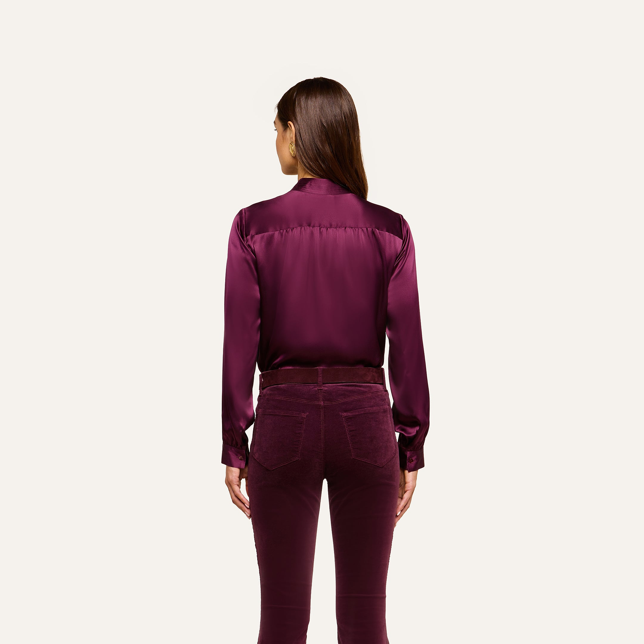 A woman stands facing away, wearing the Bianca Silk Blouse in deep purple with matching velvet pants, against a plain light background.