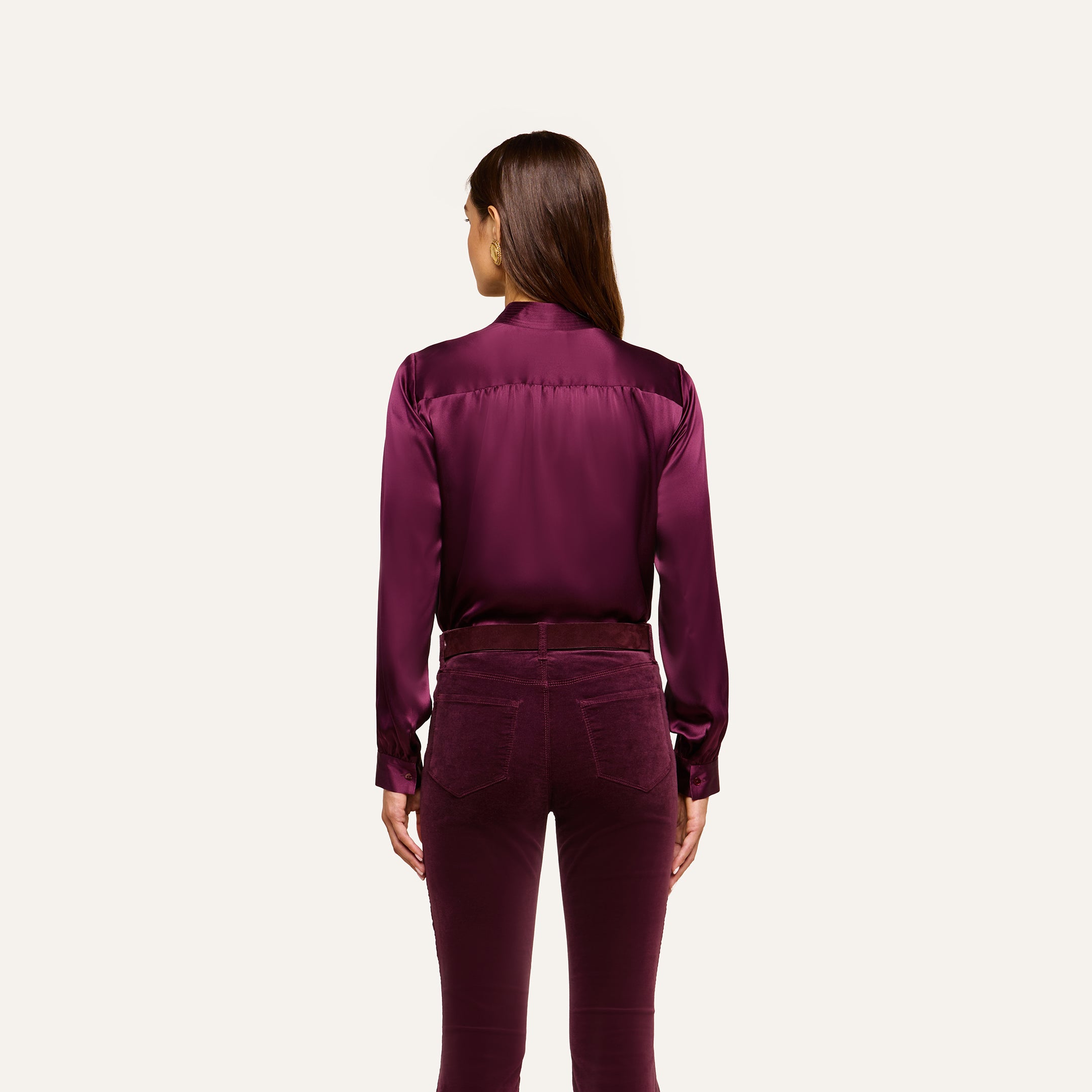 A woman stands facing away, wearing the Bianca Silk Blouse in deep purple with matching velvet pants, against a plain light background.