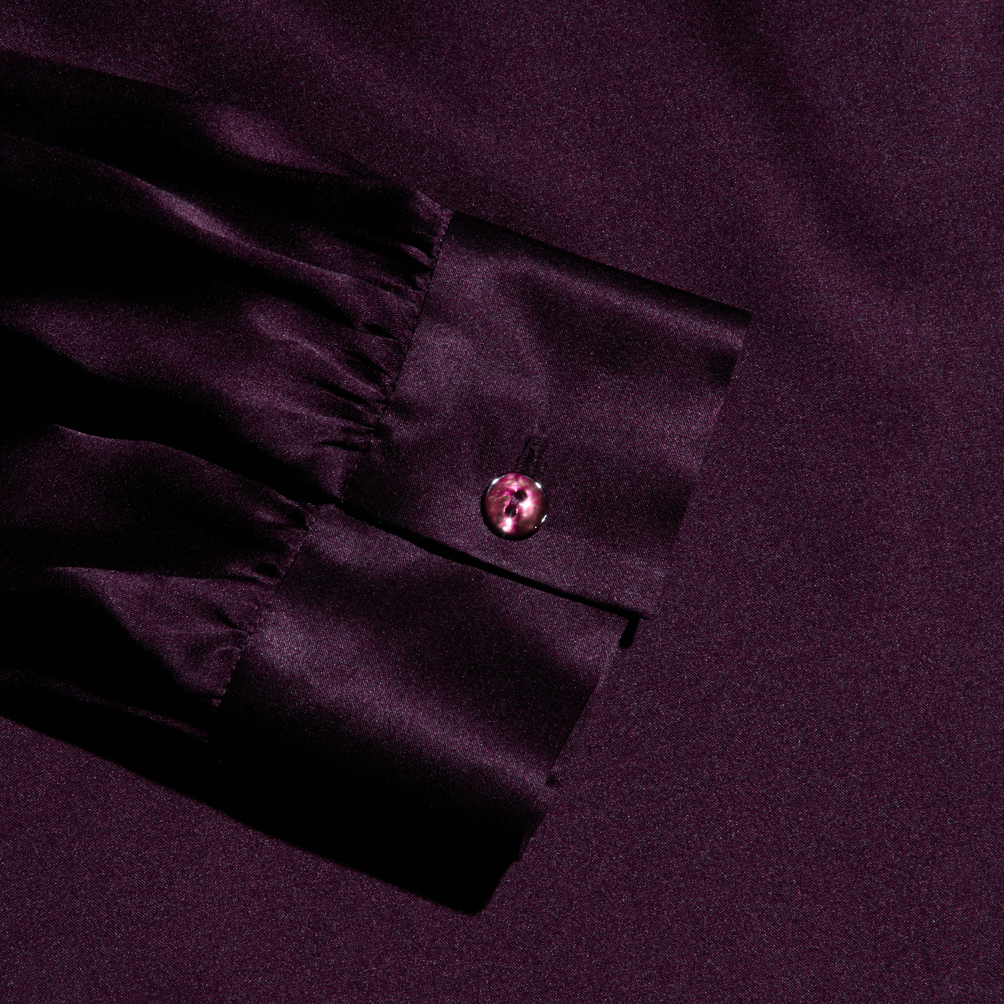 A close-up, top-down view of the Bianca Silk Blouse’s sleeve cuff in deep purple silk charmeuse. The gathered cuff is finished with a single matching button centered on the band, showcasing the blouse’s smooth, slightly reflective fabric.