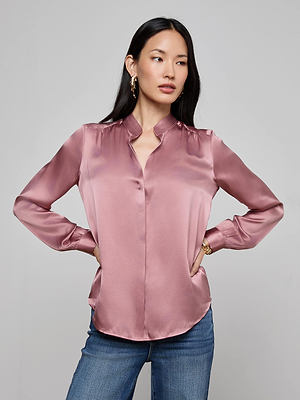 A woman with long dark hair models the L'AGENCE Bianca Silk Blouse in shiny light pink with blue jeans, standing hands on hips against a plain light gray background and gazing slightly to the side.