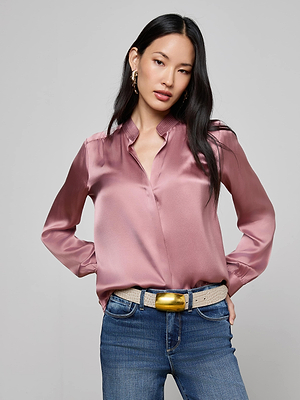 A woman with long black hair wears the Bianca Silk Blouse by L'AGENCE in shiny light pink, styled with blue jeans, a beige belt with a gold buckle, and gold earrings, standing hands-in-pockets against a plain light gray background.