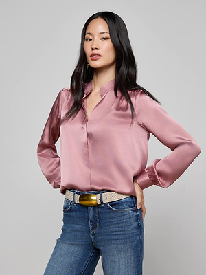 A woman with long black hair wears the L'AGENCE Bianca Silk Blouse in mauve, styled with blue jeans and a tan belt with a large gold buckle. She stands confidently, hands on hips, against a light gray background.