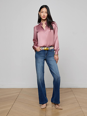 A woman with long dark hair poses confidently indoors against a plain wall, wearing the L'AGENCE Bianca Silk Blouse in light pink, blue jeans, nude heels, and a belt. One hand is in her pocket as she stands on a wooden floor.
