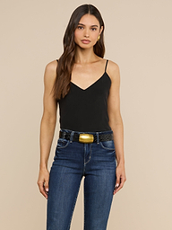 A woman with long brown hair wears the Jane Silk Camisole in black, featuring adjustable straps, paired with dark blue jeans and a wide black belt with a large gold buckle, standing against a plain light background.