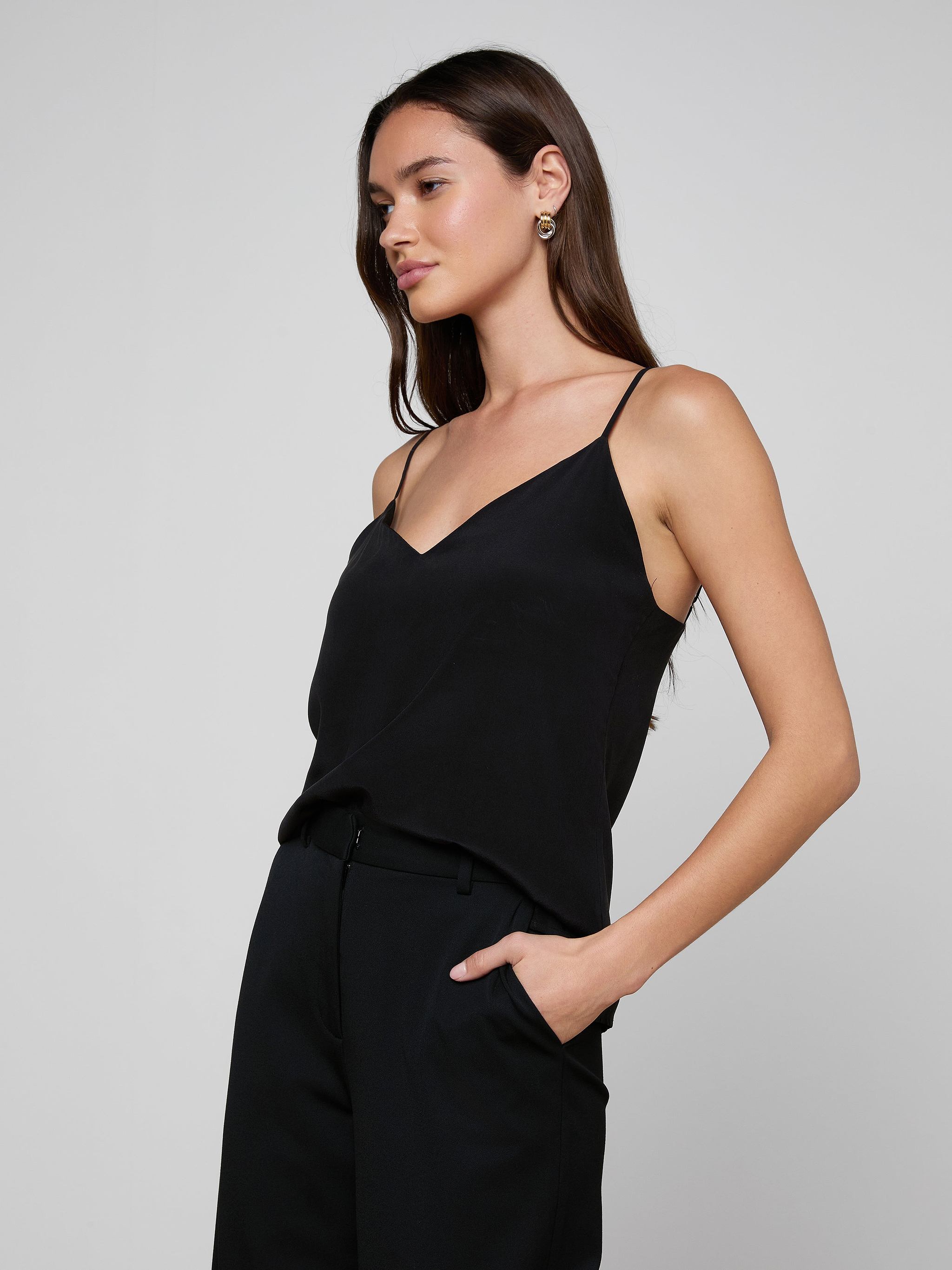 A woman wearing the Jane Silk Camisole with adjustable straps and blue jeans stands against a plain light background. She has long brown hair, gold hoop earrings, and a black belt with a large gold buckle.
