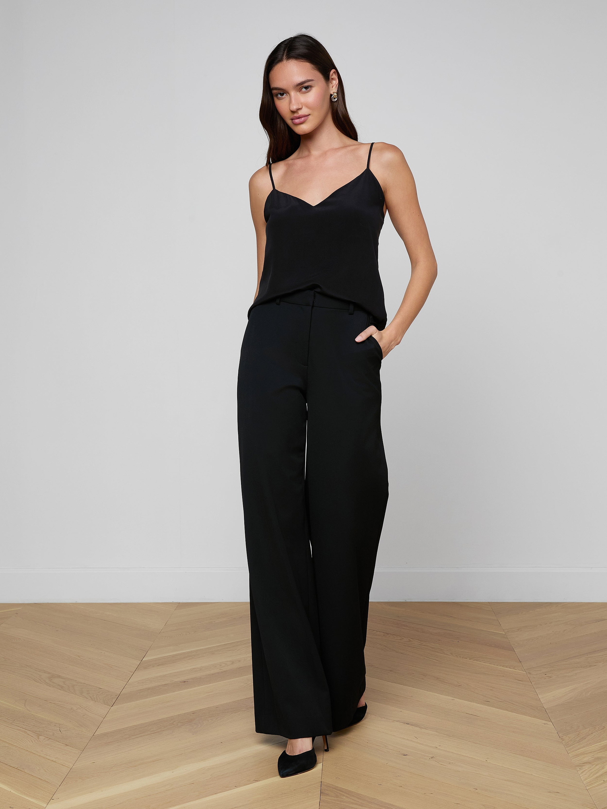 A woman with long hair and a neutral expression stands in a plain room, wearing the Jane Silk Camisole—black with adjustable straps—paired with dark blue bootcut jeans, a black belt featuring a gold buckle, and black heels.