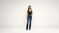 A woman stands against a plain white background, wearing a black sleeveless top, dark blue flared jeans, black heels, and a belt. She has long, straight brown hair and looks directly at the camera.