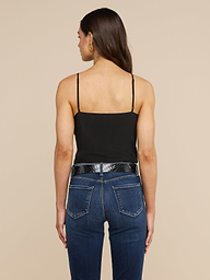 A woman with long brown hair stands facing away, wearing the Jane Silk Camisole with adjustable straps, paired with dark blue jeans, a black belt, and gold hoop earrings against a plain light background.