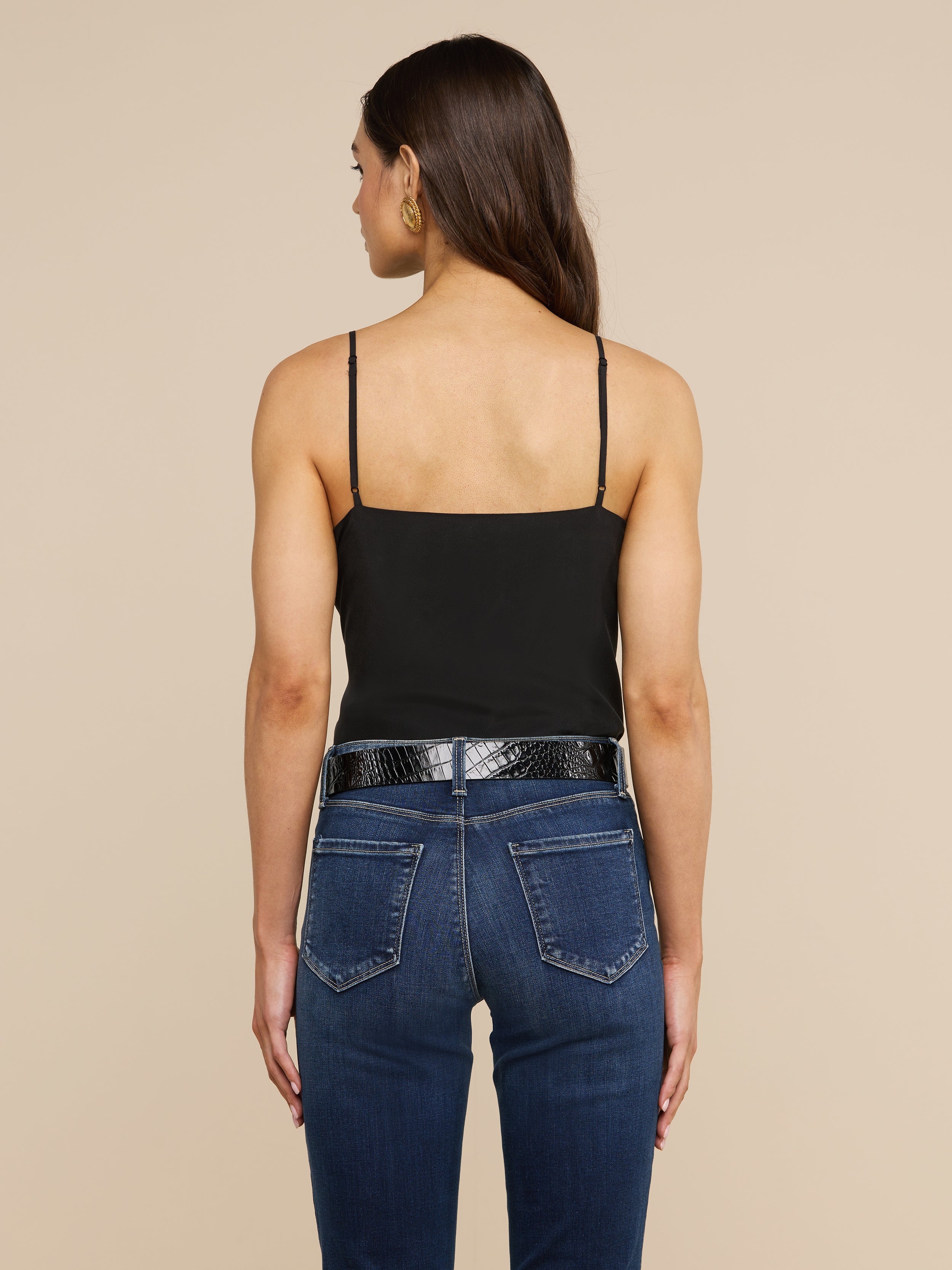 A woman with long brown hair stands facing away, wearing the Jane Silk Camisole with adjustable straps, paired with dark blue jeans, a black belt, and gold hoop earrings against a plain light background.