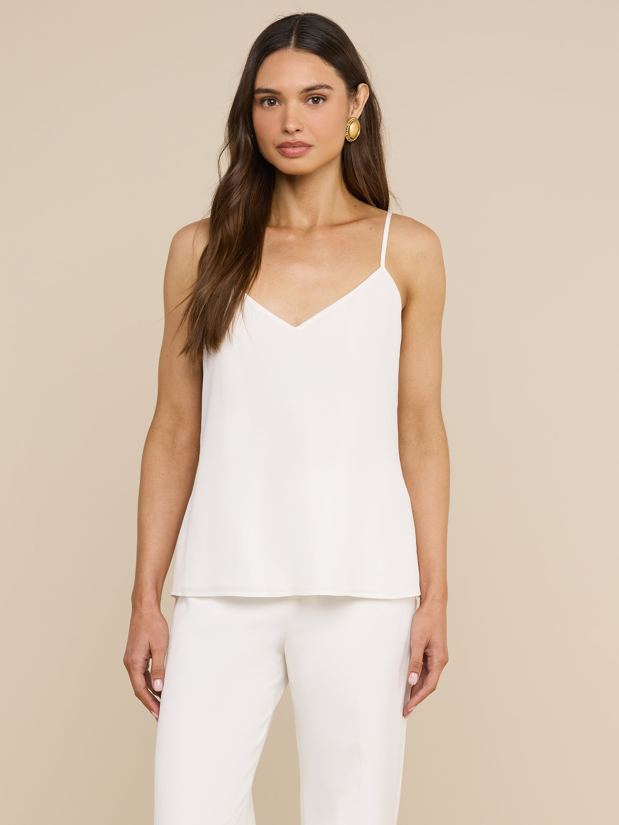 A woman with long brown hair wears the Jane Silk Camisole, featuring adjustable straps, paired with matching white pants. She faces forward against a plain light background and accessorizes with gold hoop earrings.