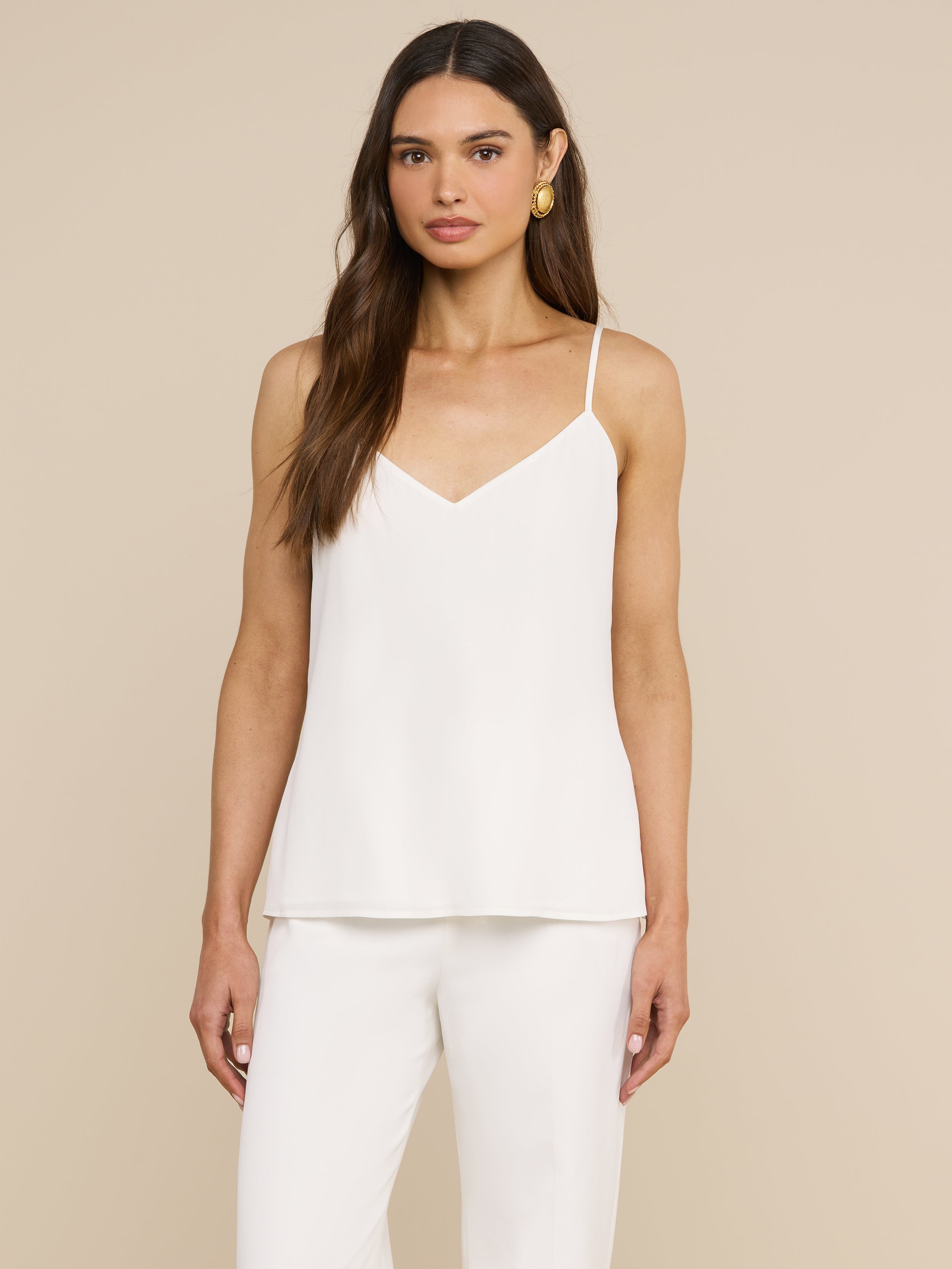 A woman with long brown hair wears the Jane Silk Camisole, featuring adjustable straps, paired with matching white pants. She faces forward against a plain light background and accessorizes with gold hoop earrings.