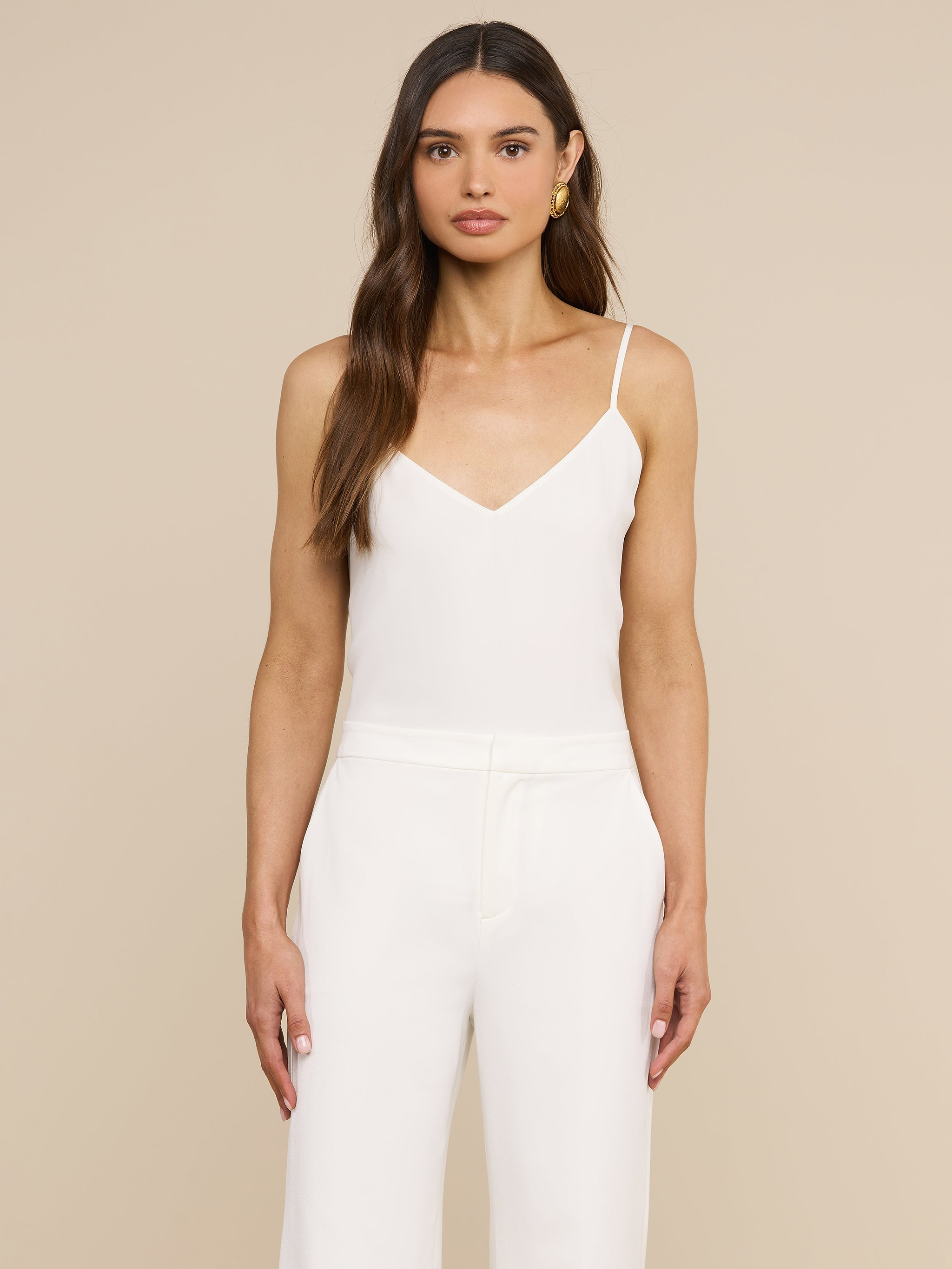A woman with long brown hair wears the Jane Silk Camisole, a sleeveless white top with adjustable straps, paired with high-waisted white pants. She stands against a plain light background, facing forward with a neutral expression.