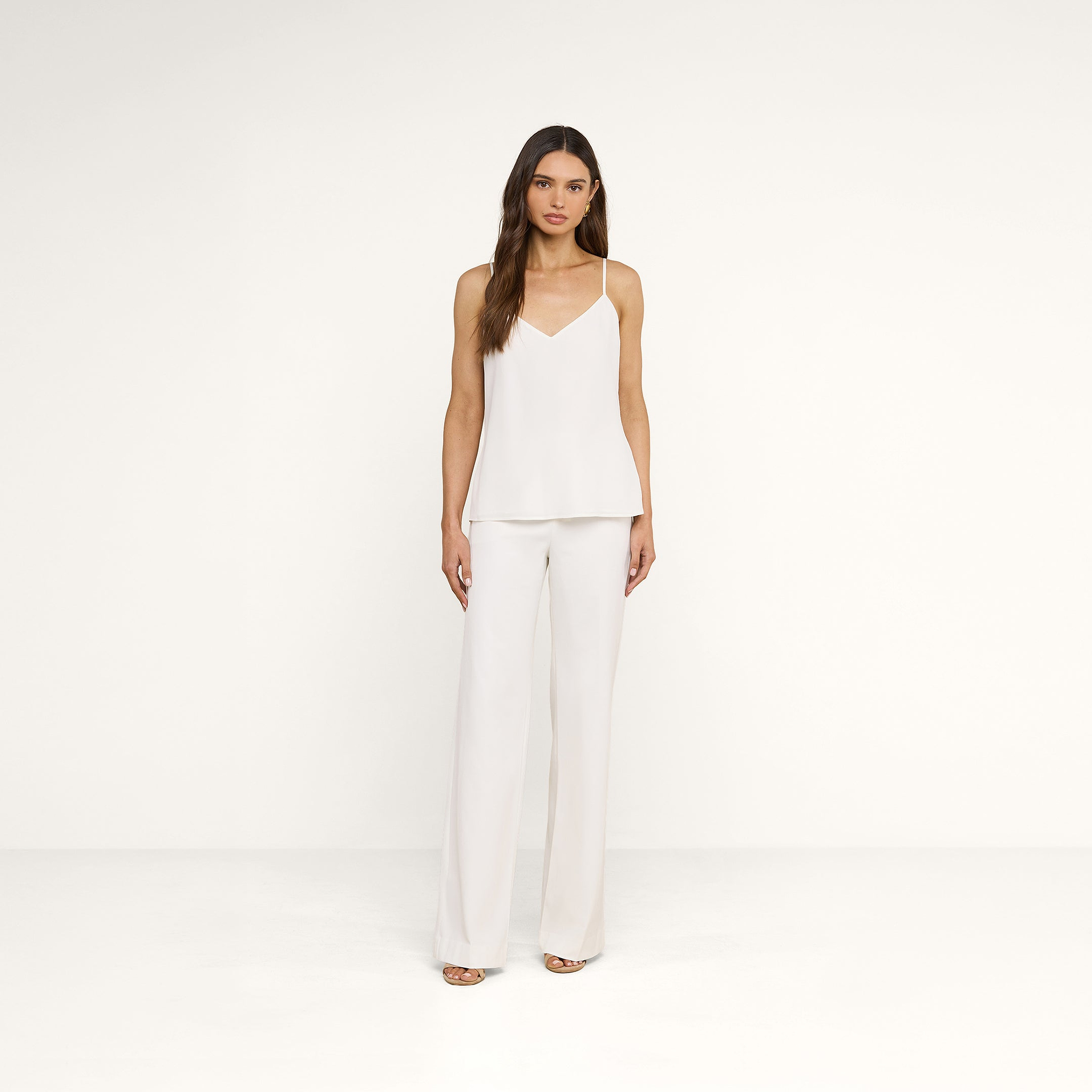 A woman with long brown hair wears the Jane Silk Camisole—a bias-cut silk top with adjustable straps—paired with white wide-leg pants, standing against a plain white background with a neutral expression.