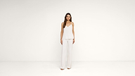 A woman stands against a plain white background, wearing a sleeveless white top and wide-leg white pants, with loose hair and neutral expression.