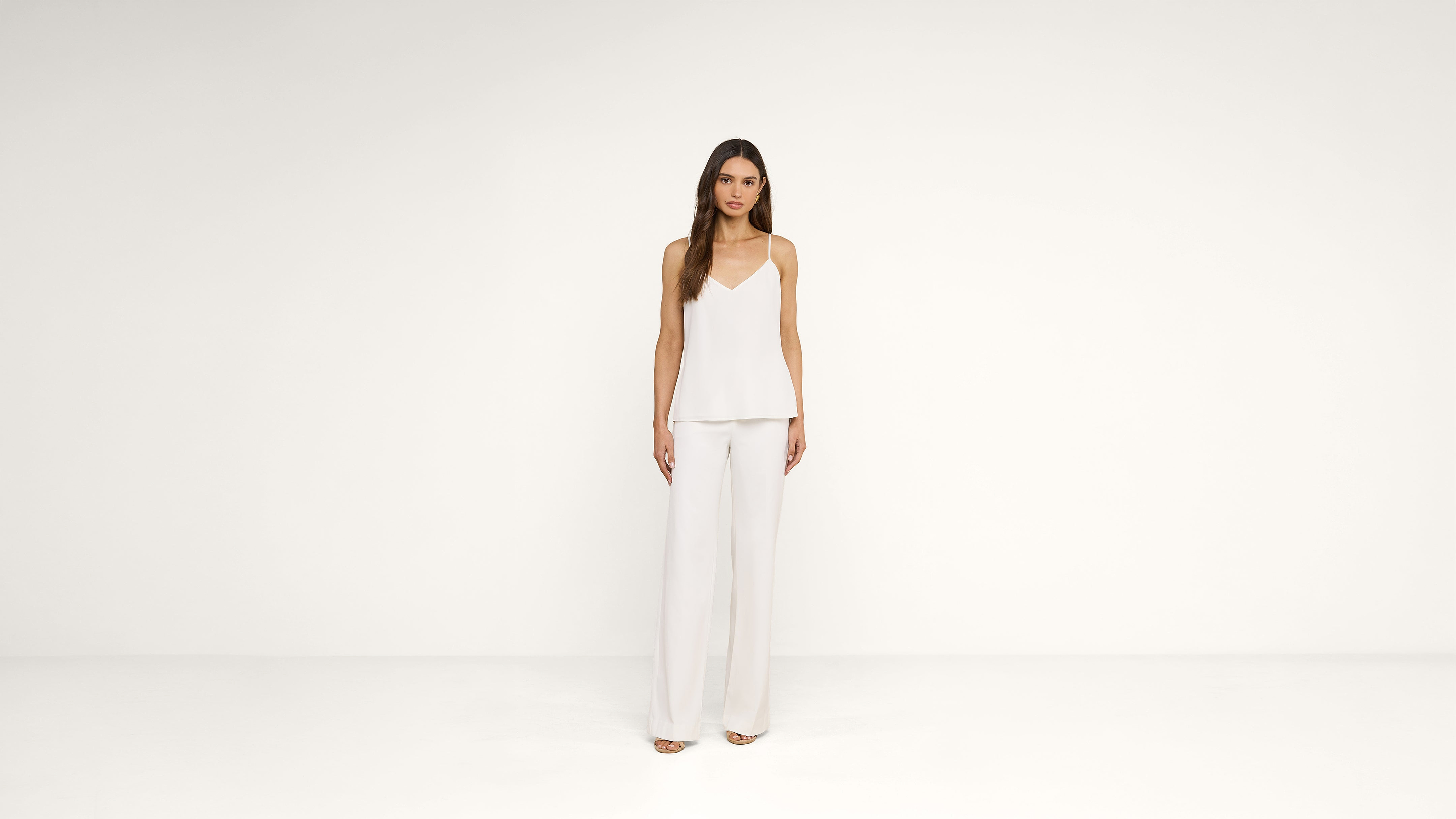 A woman stands against a plain white background, wearing a sleeveless white top and wide-leg white pants, with loose hair and neutral expression.