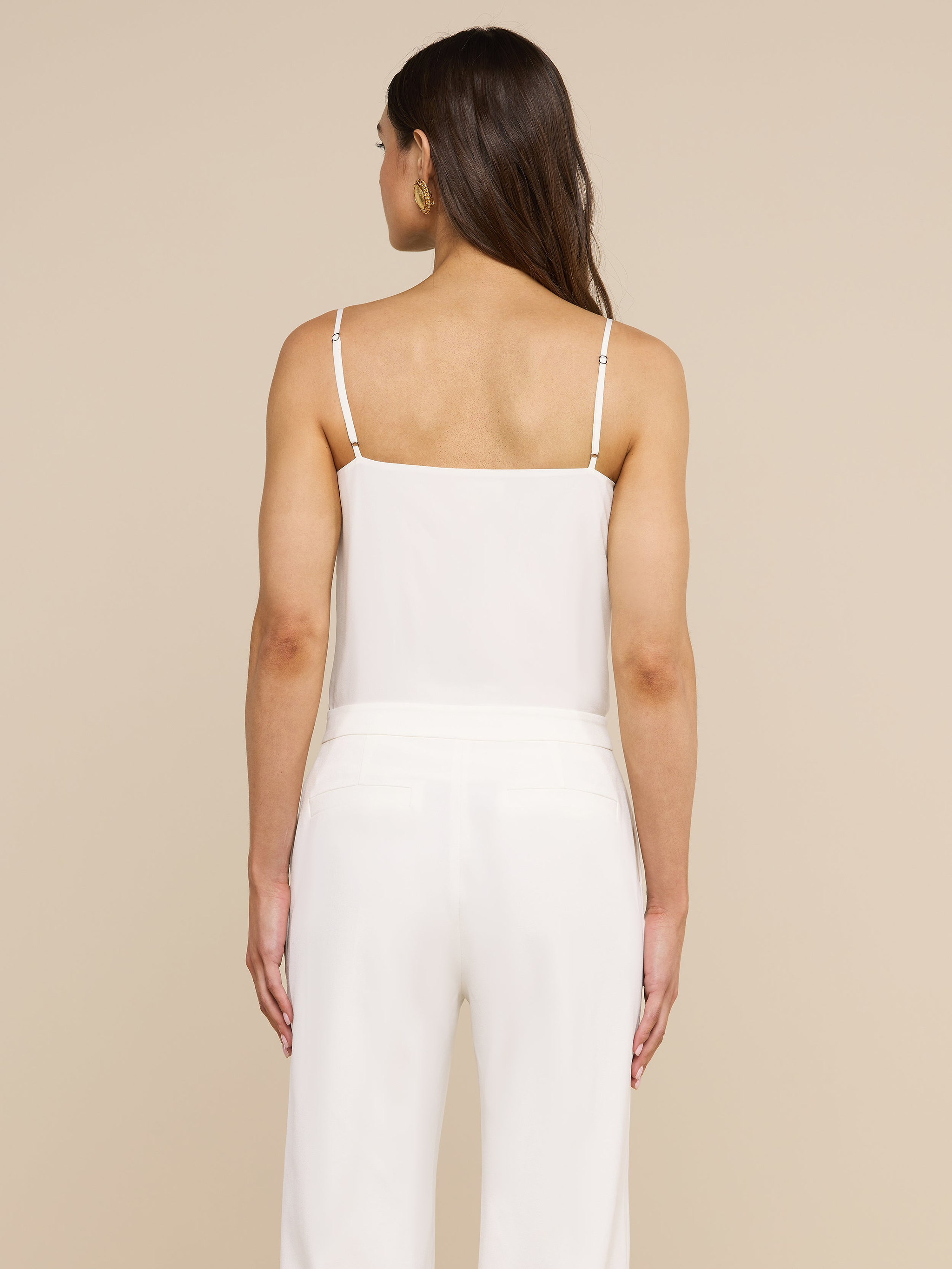 A woman with long brown hair stands facing away against a plain light background, wearing the Jane Silk Camisole—a bias-cut silk top with adjustable straps—paired with white pants.