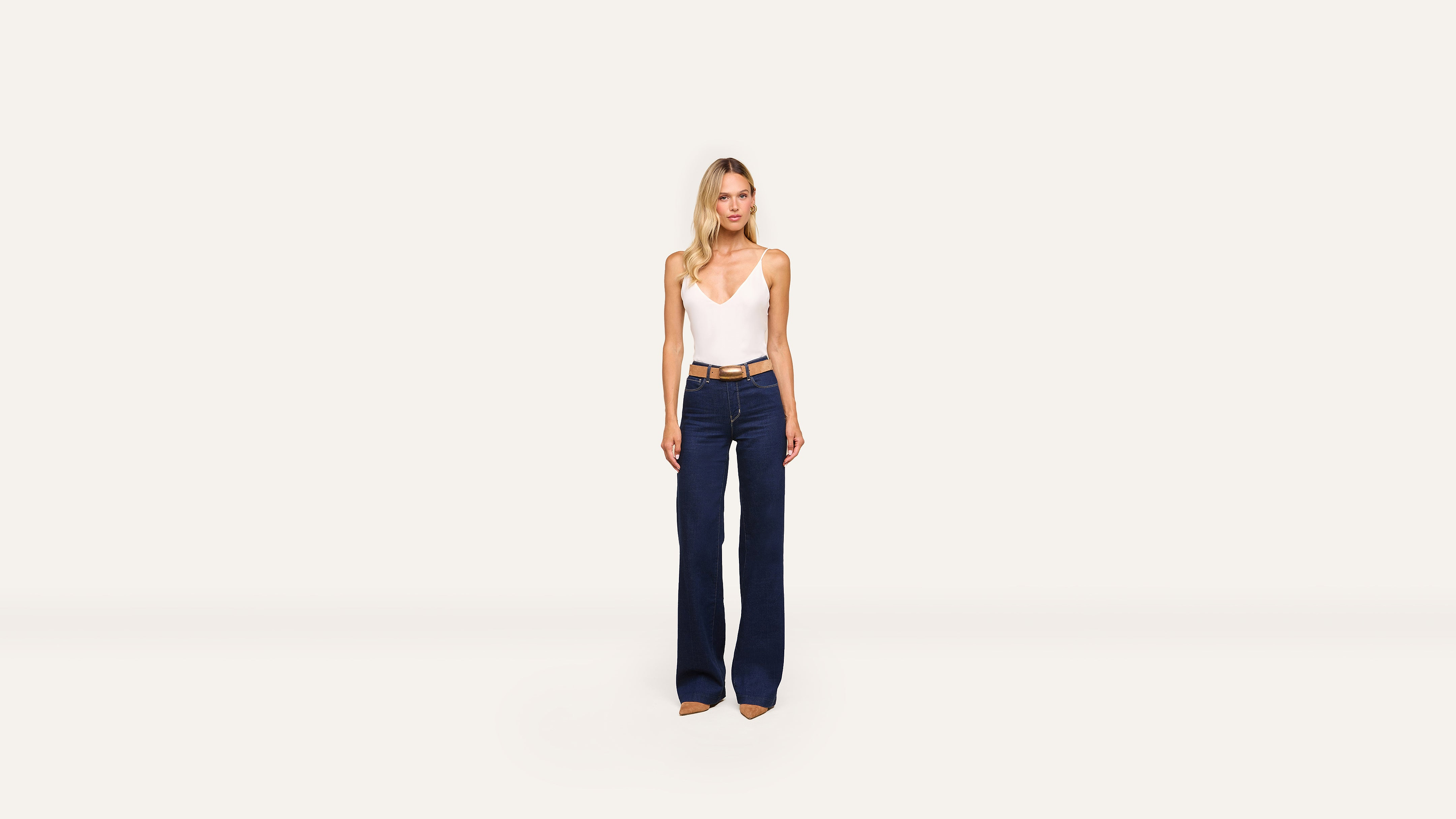 A woman stands against a plain white background, wearing a white sleeveless top, wide-leg dark blue jeans, and stylish tan heels.