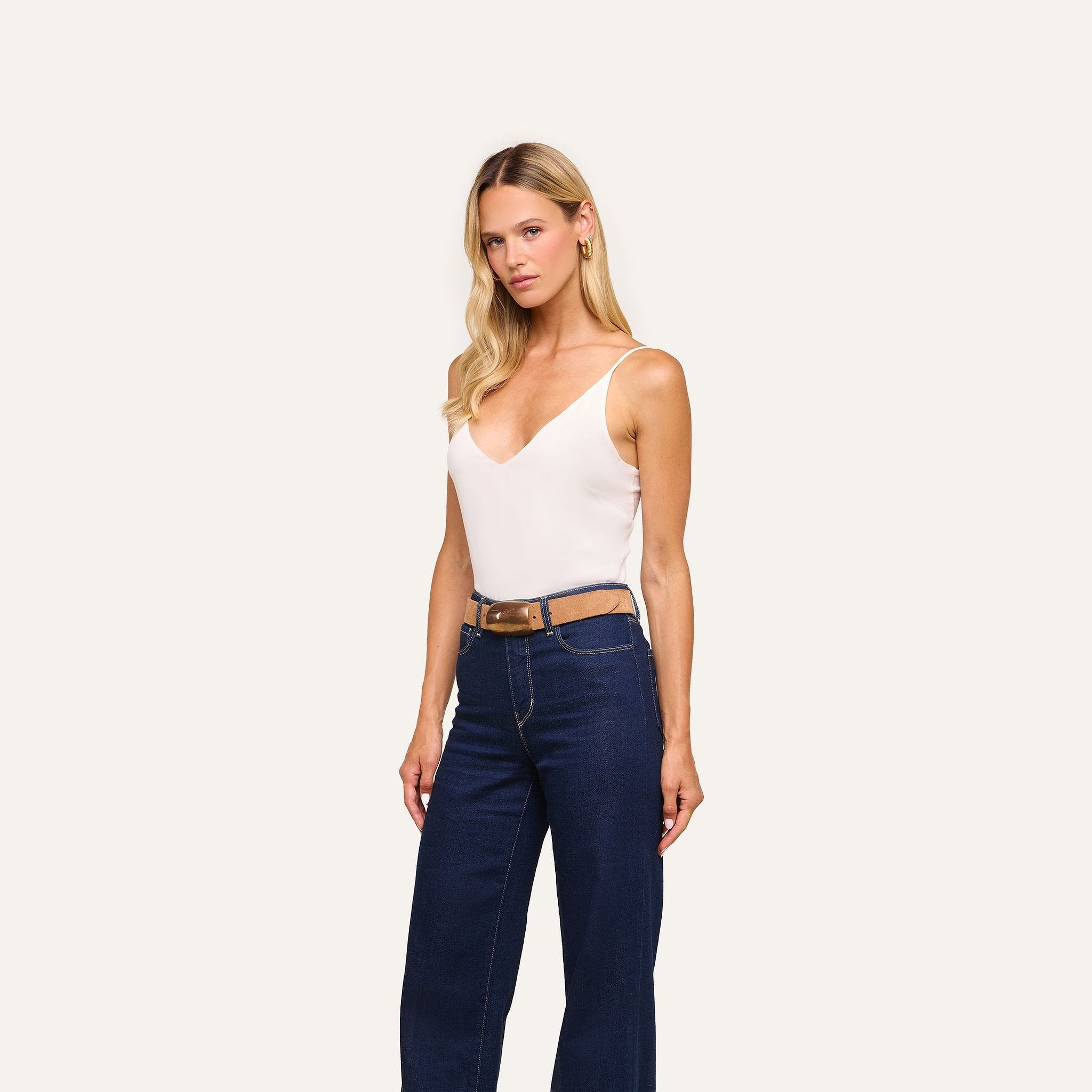 Woman with long blonde hair wears the Gabriella Silk Camisole, featuring adjustable straps, paired with blue high-waisted jeans and a brown belt, standing against an off-white background.