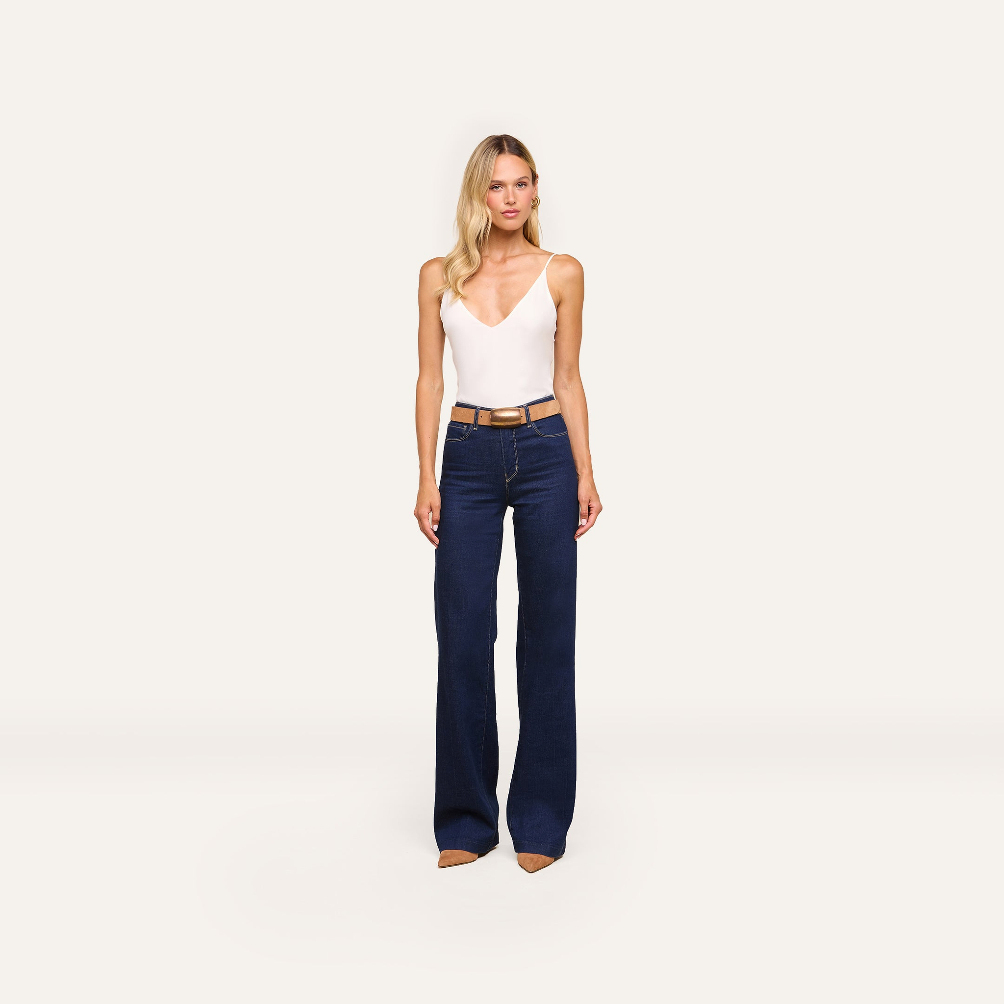 A woman faces forward wearing the Gabriella Silk Camisole in ivory with adjustable straps, paired with dark blue high-waisted wide-leg jeans, a brown belt, and brown pointed shoes against a plain light background.
