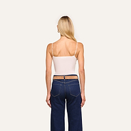 A woman with long blonde hair faces away, wearing the Gabriella Silk Camisole with adjustable straps and dark blue jeans with cutout waist details, against a plain light background.