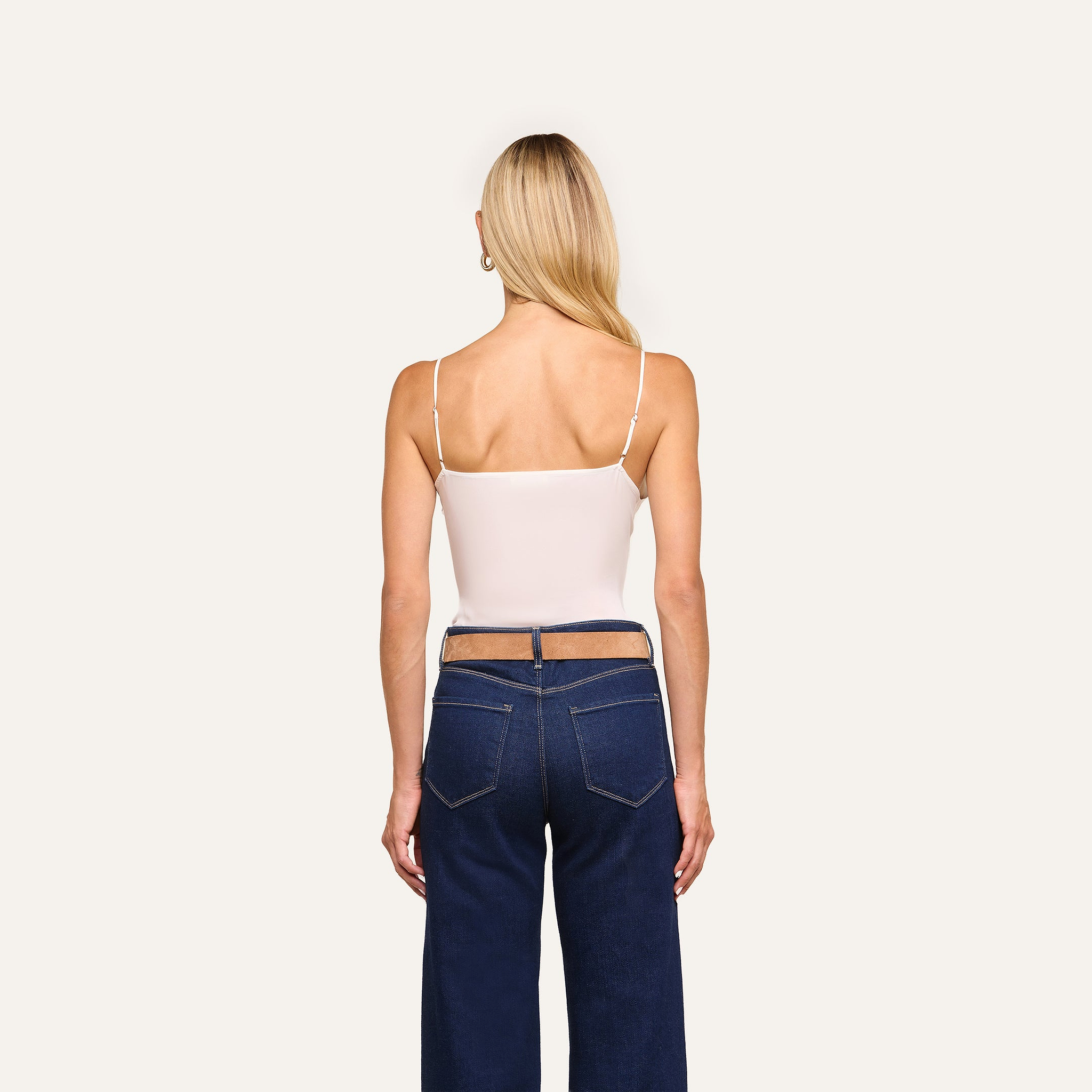 A woman with long blonde hair faces away, wearing the Gabriella Silk Camisole with adjustable straps and dark blue jeans with cutout waist details, against a plain light background.