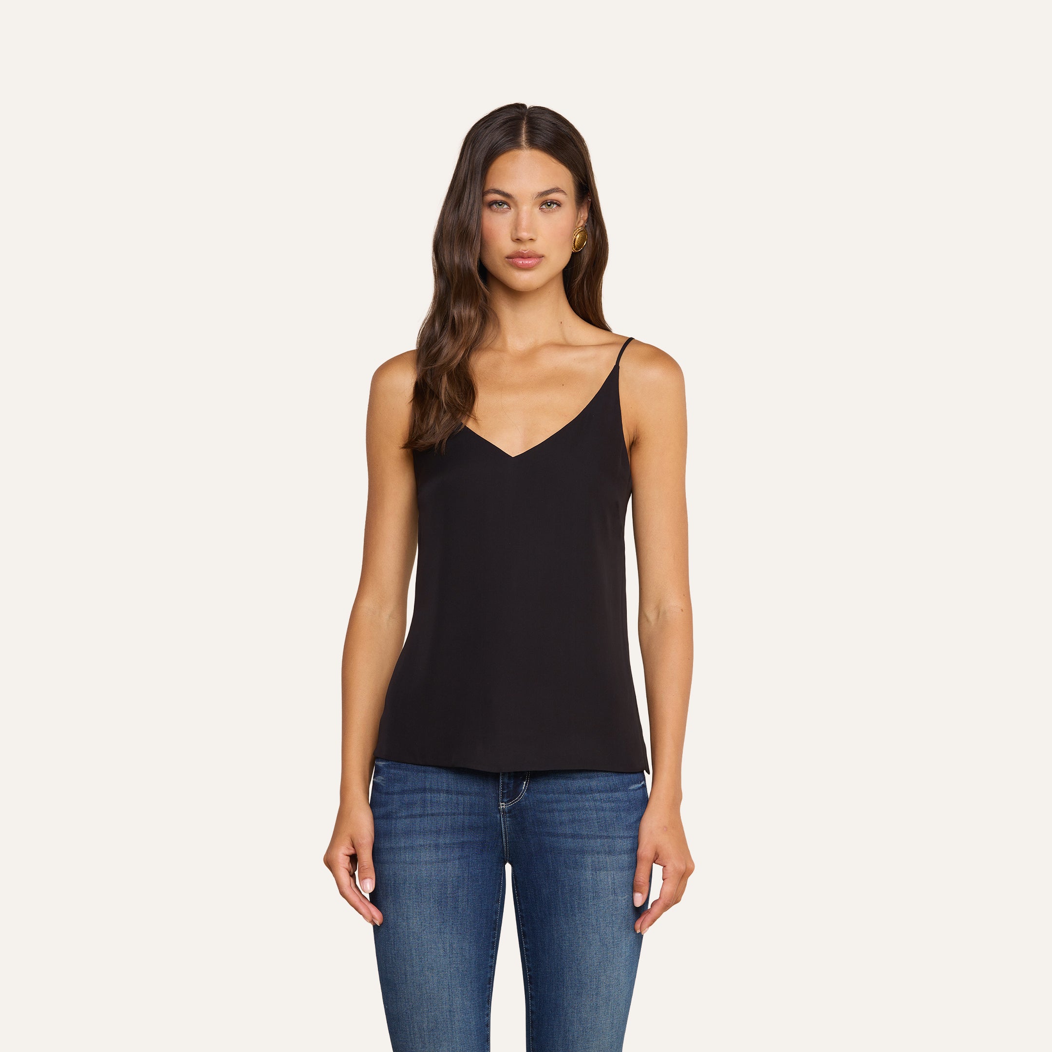 Woman with long brown hair wearing the Gabriella Silk Camisole in black and blue jeans, standing against a plain light background, facing forward with her arms relaxed by her sides. This versatile camisole adds effortless elegance to her look.