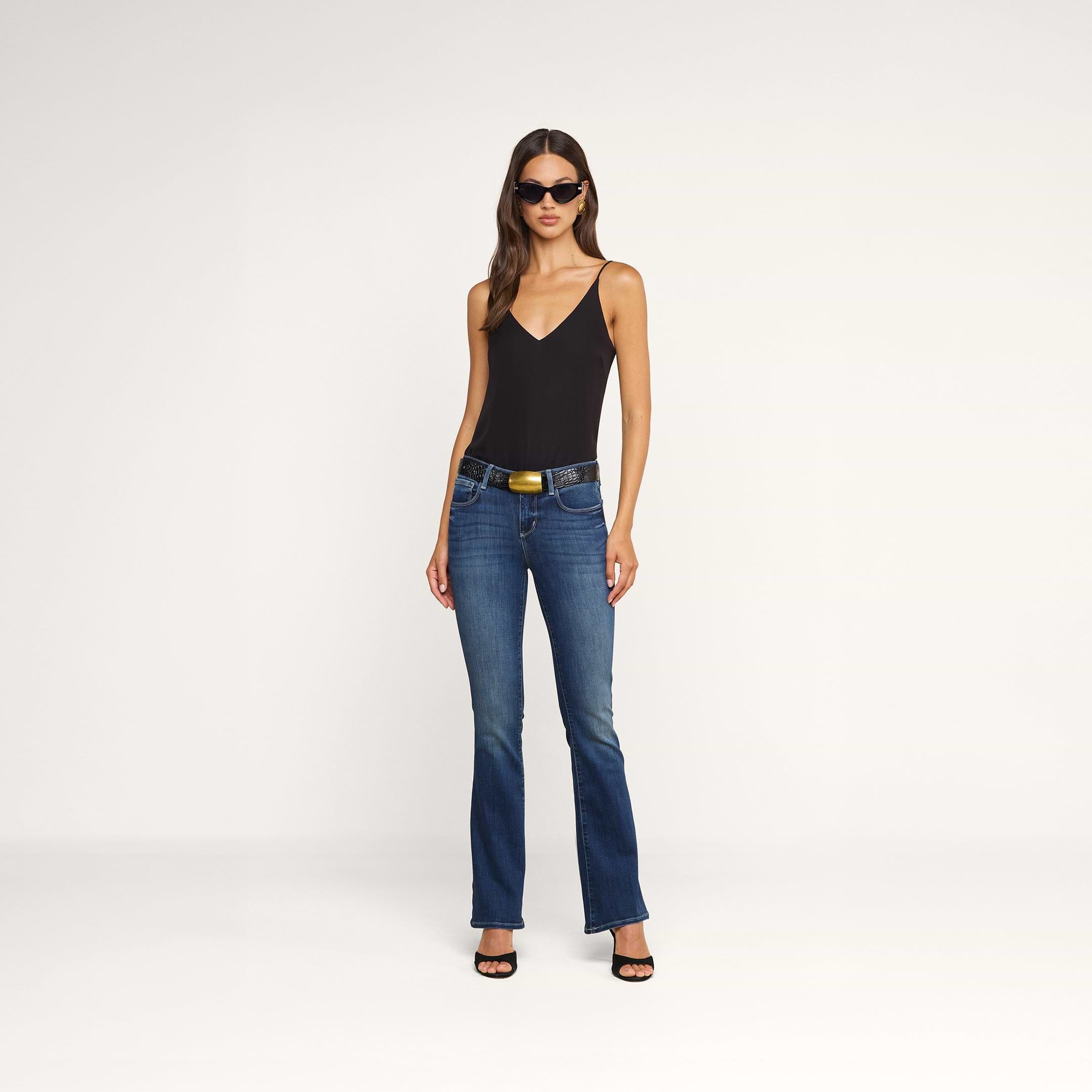 A woman stands confidently against a plain white background, wearing sunglasses, blue flared jeans, black heels, and the Gabriella Silk Camisole.