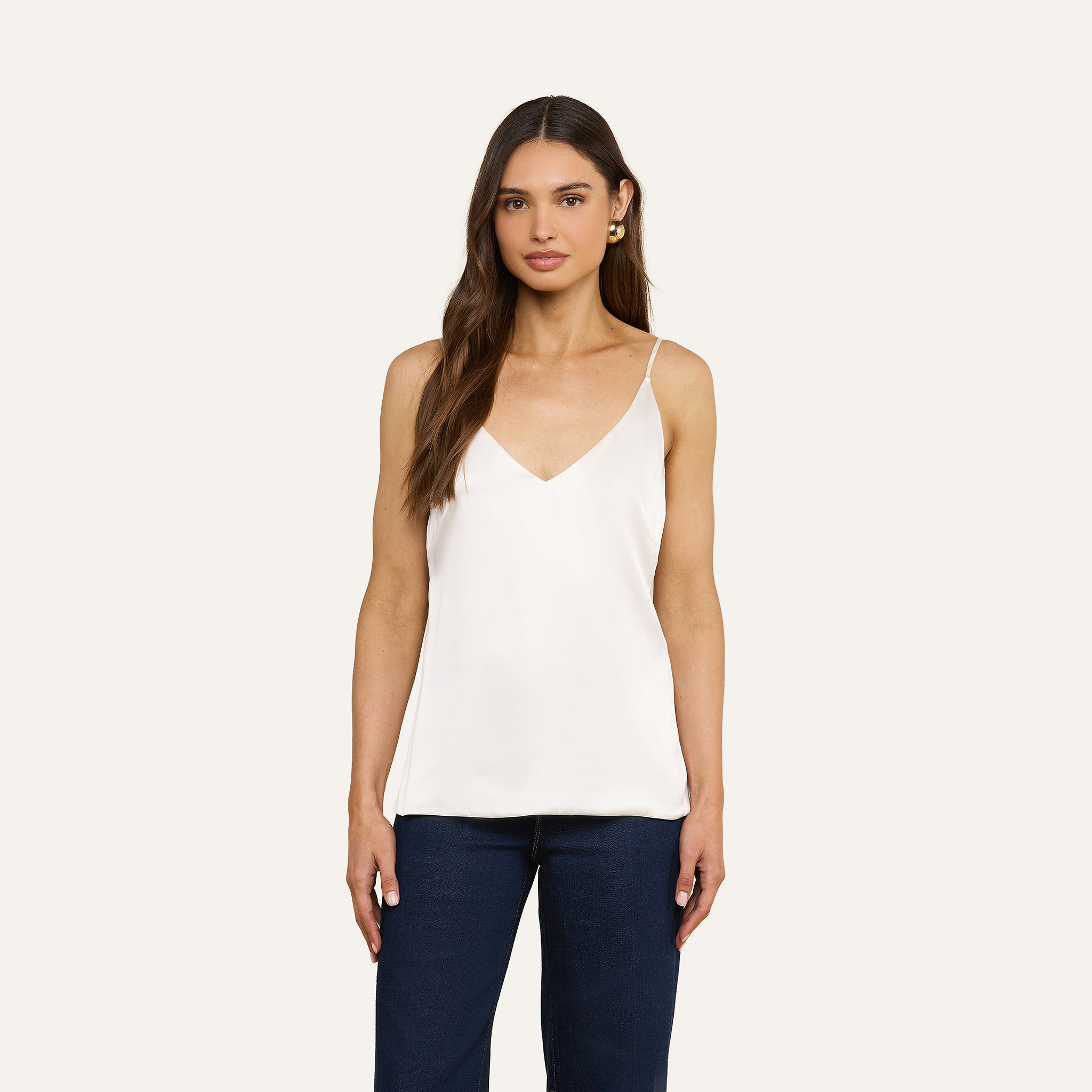 A woman with long brown hair wears the Gabriella Silk Camisole, featuring adjustable spaghetti straps, paired with dark blue jeans against a plain beige background.