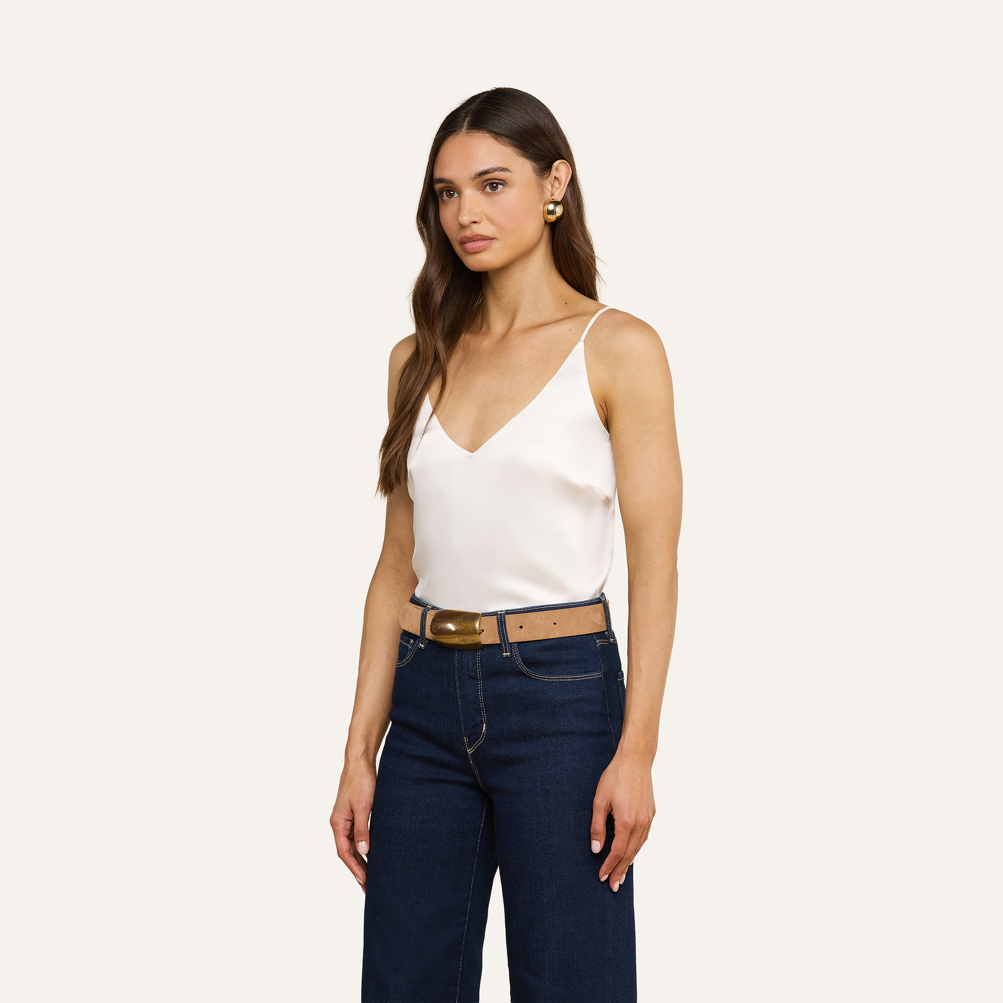 A woman with long brown hair, wearing the Gabriella Silk Camisole with adjustable spaghetti straps, dark blue jeans, a brown belt with a gold buckle, and gold earrings, stands against a beige background.