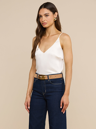 A woman with long brown hair, wearing the Gabriella Silk Camisole with adjustable spaghetti straps, dark blue jeans, a brown belt with a gold buckle, and gold earrings, stands against a beige background.