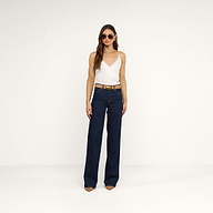 Una donna in occhiali da sole è in posa su uno sfondo beige, indossando la canotta Gabriella Silk bianca, senza maniche e con scollo a V, jeans blu scuro a vita alta e a gamba larga, una cintura marrone e scarpe marroni a punta, per un look chic a strati.