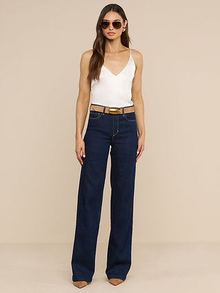 Una donna in occhiali da sole è in posa su uno sfondo beige, indossando la canotta Gabriella Silk bianca, senza maniche e con scollo a V, jeans blu scuro a vita alta e a gamba larga, una cintura marrone e scarpe marroni a punta, per un look chic a strati.