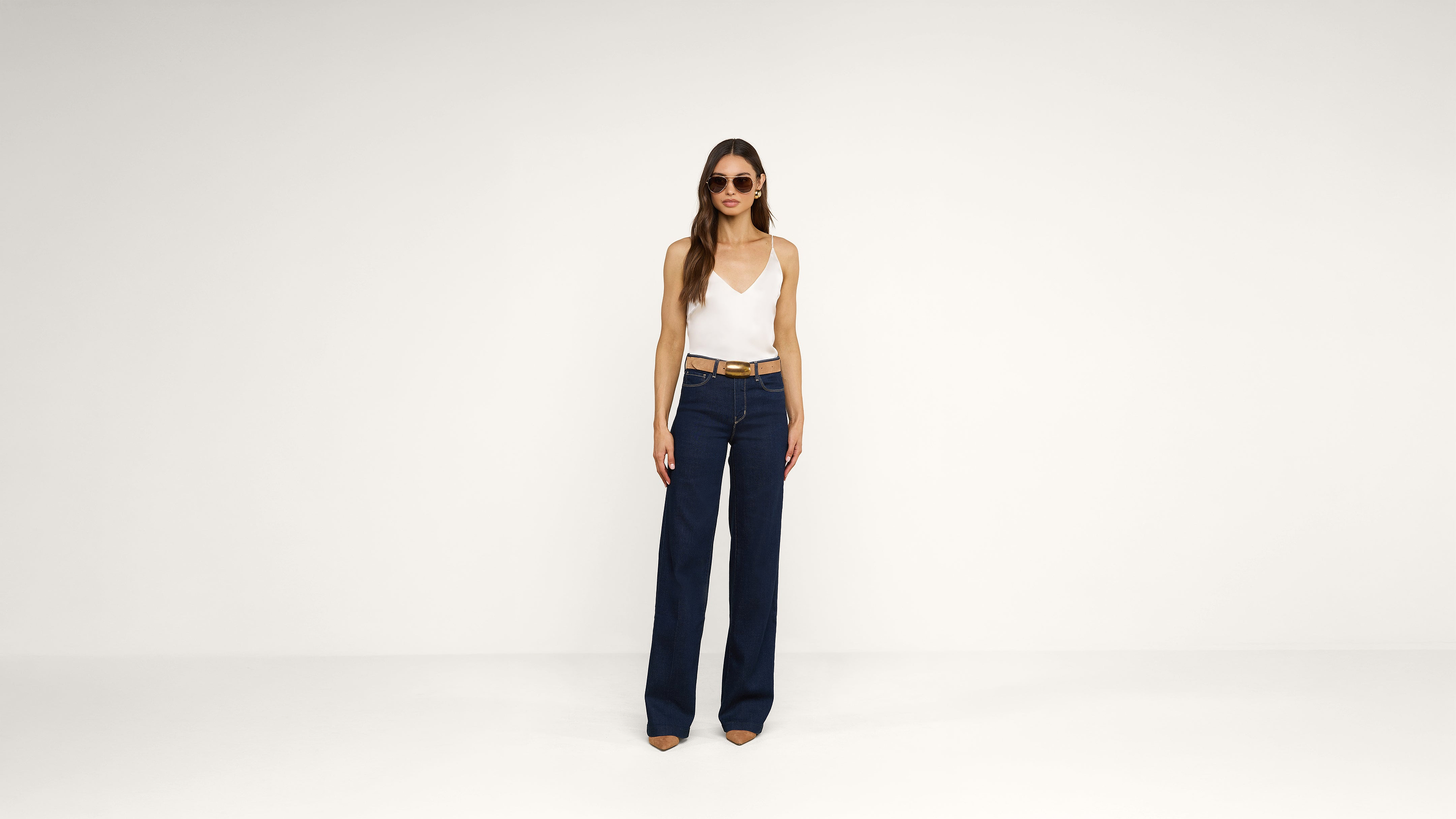 Woman wearing sunglasses, a white sleeveless top, a brown belt, and high-waisted wide-leg blue jeans stands against a plain white background.