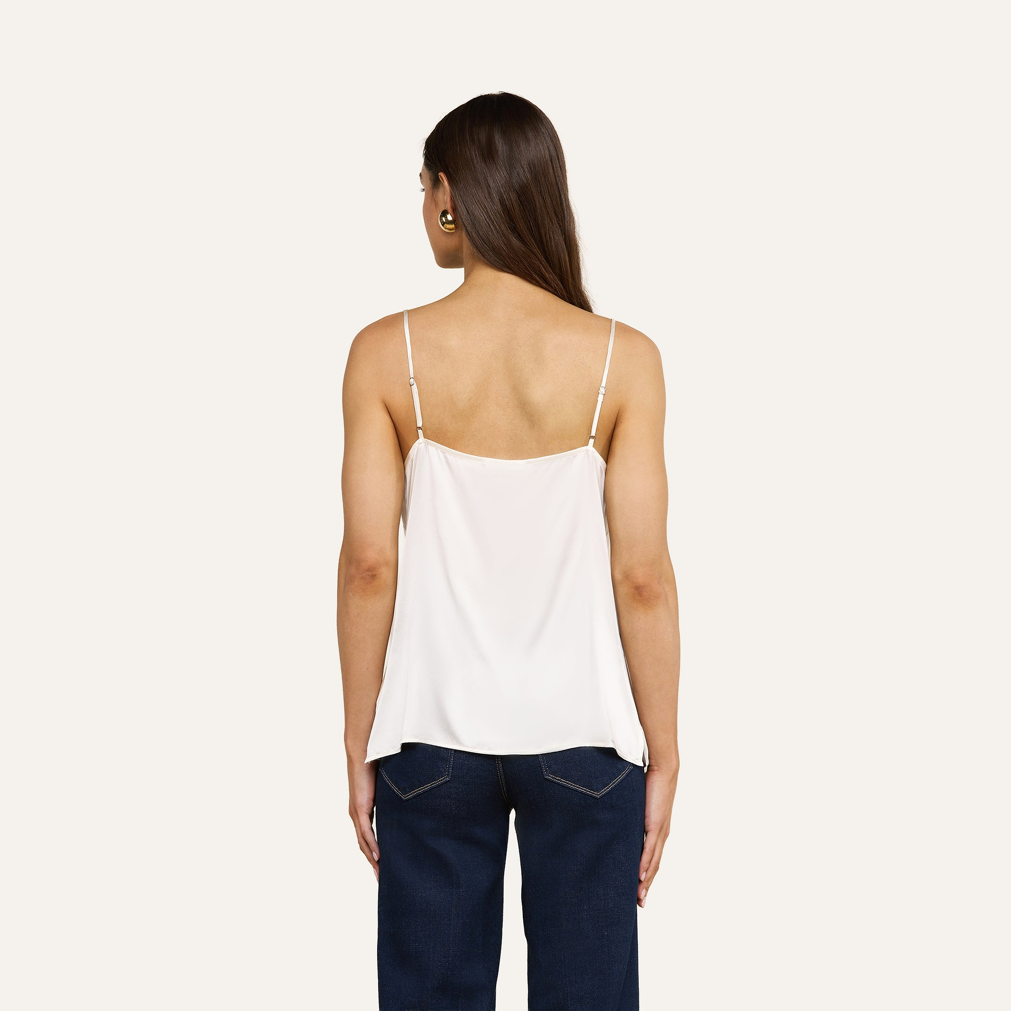 Eine Frau mit langen braunen Haaren steht mit dem Rücken zur Kamera vor einem beigen Hintergrund und trägt das „Gabriella Silk Camisole“ – ein weißes Top mit verstellbaren Spaghettiträgern – sowie eine dunkelblaue Jeans.