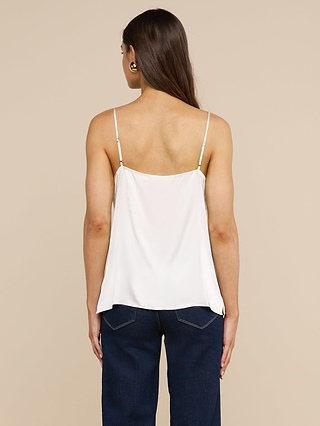 Eine Frau mit langen braunen Haaren steht mit dem Rücken zur Kamera vor einem beigen Hintergrund und trägt das „Gabriella Silk Camisole“ – ein weißes Top mit verstellbaren Spaghettiträgern – sowie eine dunkelblaue Jeans.
