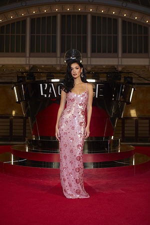 A woman stands on a red carpet in a sleeveless, floor-length pink sequin gown. She has long dark hair, red lipstick, and is posing in front of a display with L’AGENCE written on it.