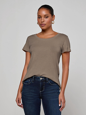 A woman wearing the L'AGENCE Cory Cotton Scoopneck Tee in taupe and dark blue jeans stands against a plain light gray background, gazing at the camera with a neutral expression. Her hair is pulled back and she has on hoop earrings.