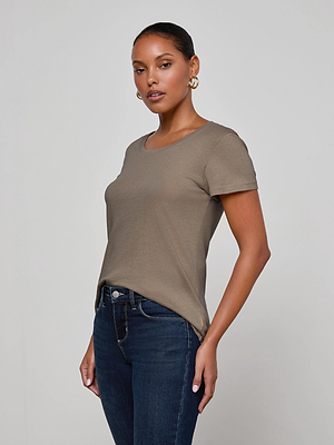 A woman with slicked-back hair wears gold hoop earrings, dark blue jeans, and the L'AGENCE Cory Cotton Scoopneck Tee in taupe. She stands against a plain light gray background, facing slightly to the side with a neutral expression.
