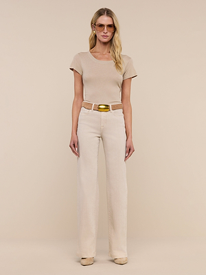 A woman with long blonde hair and a relaxed expression stands against a beige background wearing the Cory Cotton Scoopneck Tee in taupe, high-waisted light beige pants with a brown belt, beige shoes, sunglasses, and earrings.