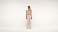 A woman wearing beige sunglasses, a beige short-sleeve top, matching wide-leg pants, a brown belt, and beige heels stands against a plain white background.