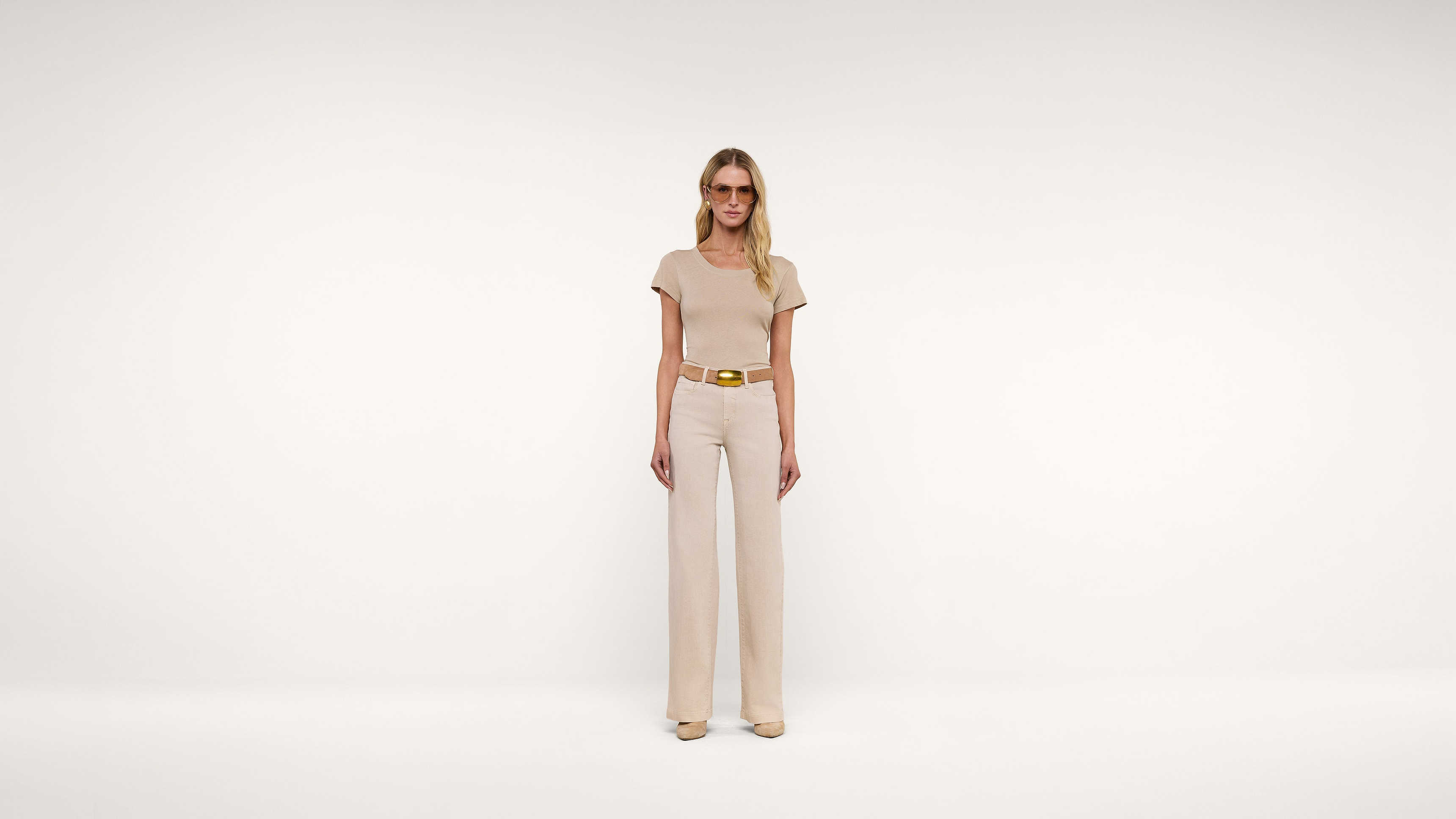 A woman wearing beige sunglasses, a beige short-sleeve top, matching wide-leg pants, a brown belt, and beige heels stands against a plain white background.