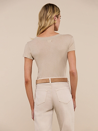 A woman with blonde hair stands facing away, wearing the Cory Cotton Scoopneck Tee in taupe and high-waisted light jeans with a brown belt, set against a neutral background.