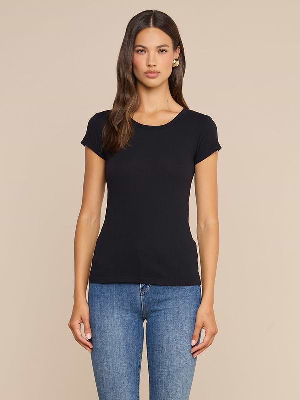 Una donna dai lunghi capelli castani indossa la maglietta Cory Cotton Scoopneck Tee in nero e jeans blu; è in piedi su uno sfondo beige a tinta unita, con lo sguardo rivolto in avanti e un’espressione neutra.