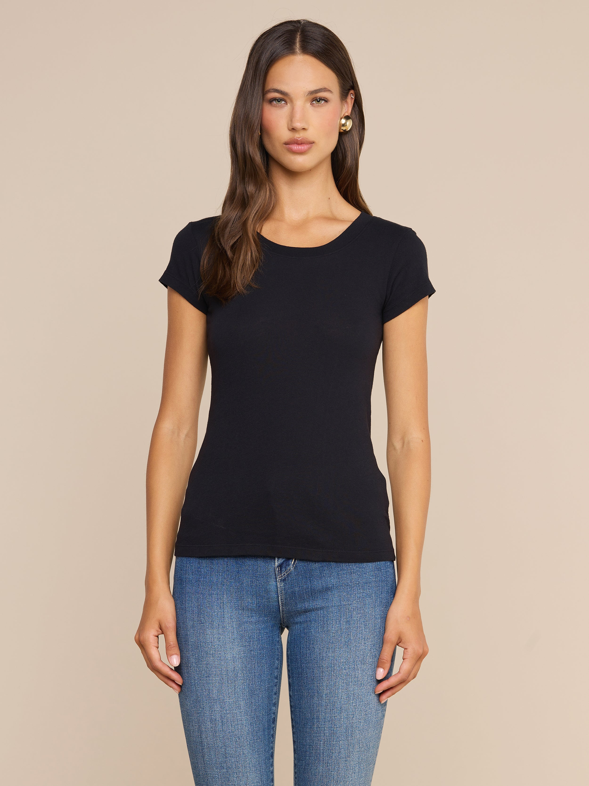 Una donna dai lunghi capelli castani indossa la maglietta Cory Cotton Scoopneck Tee in nero e jeans blu; è in piedi su uno sfondo beige a tinta unita, con lo sguardo rivolto in avanti e un’espressione neutra.