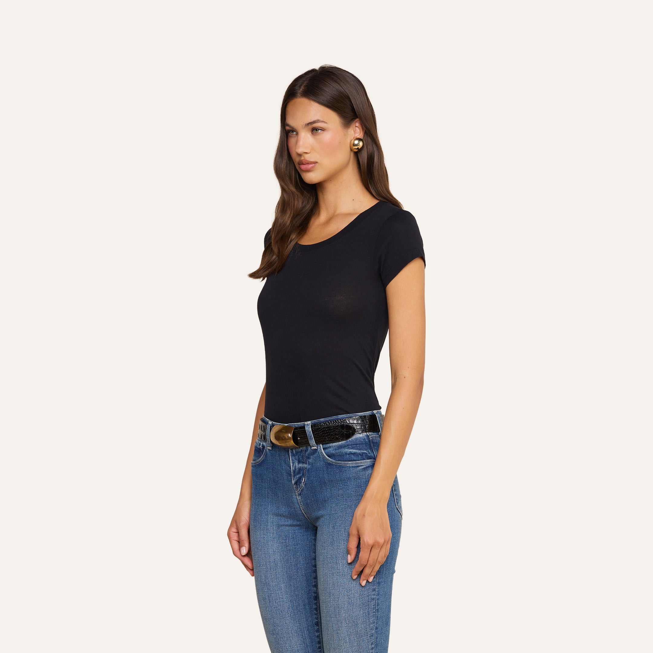 Una donna dai lunghi capelli castani è in piedi su uno sfondo beige e indossa una maglietta con scollo rotondo di Cory Cotton, jeans blu, una cintura nera con fibbia dorata e orecchini dorati.