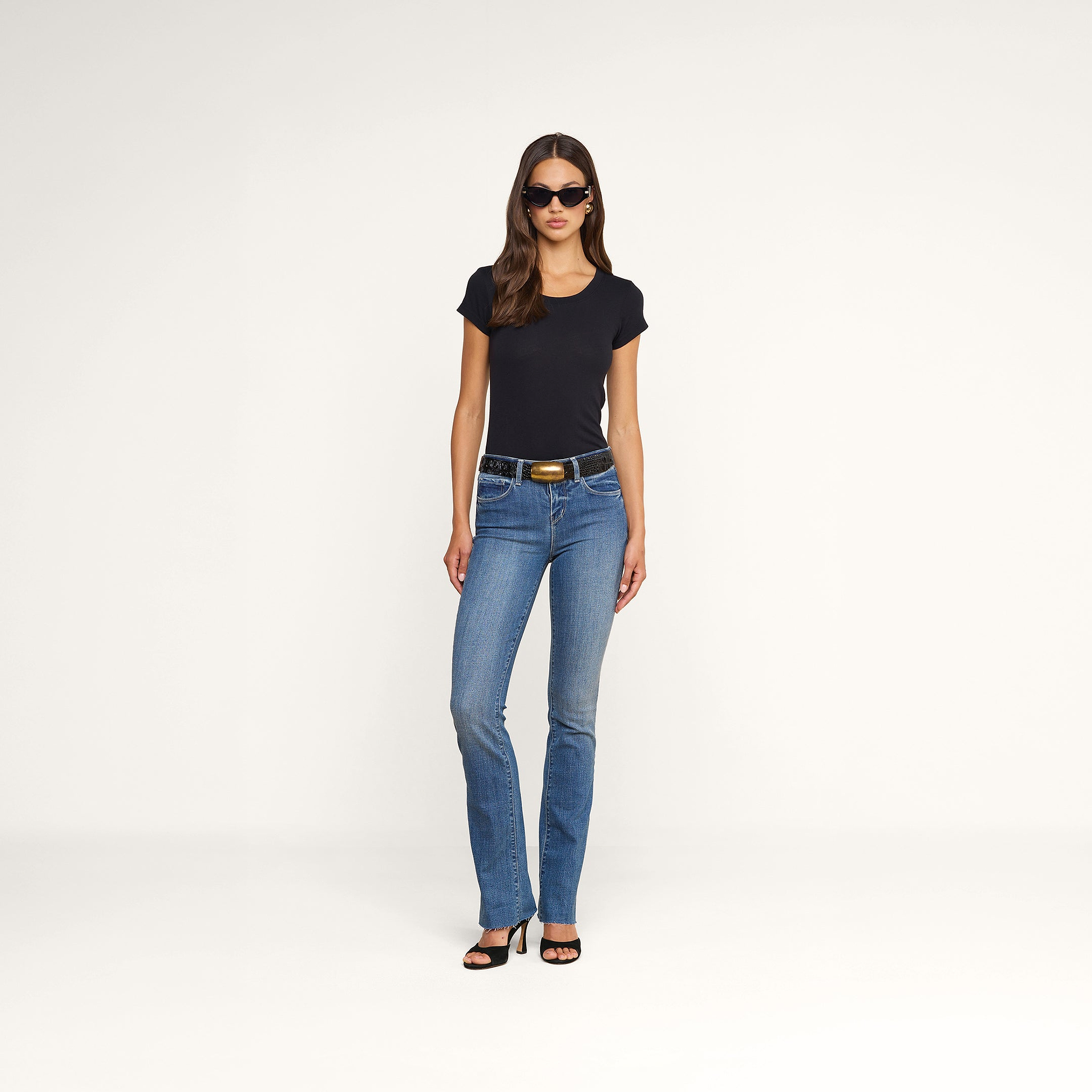 Una donna indossa occhiali da sole neri, la maglietta Cory Cotton Scoopneck Tee nera, jeans blu con cintura e sandali neri con tacco alto, in piedi su uno sfondo beige.